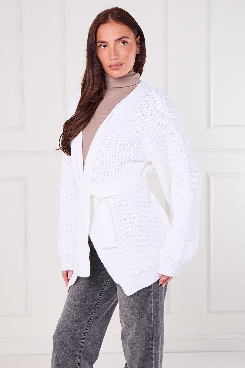 Woman wearing a white ribbed knit cardigan over a beige sweater with gray pants against a white background