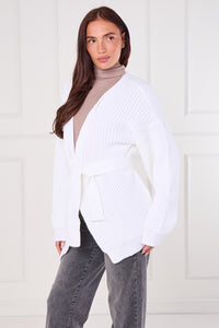 Woman wearing a white ribbed knit cardigan over a beige sweater with gray pants against a white background