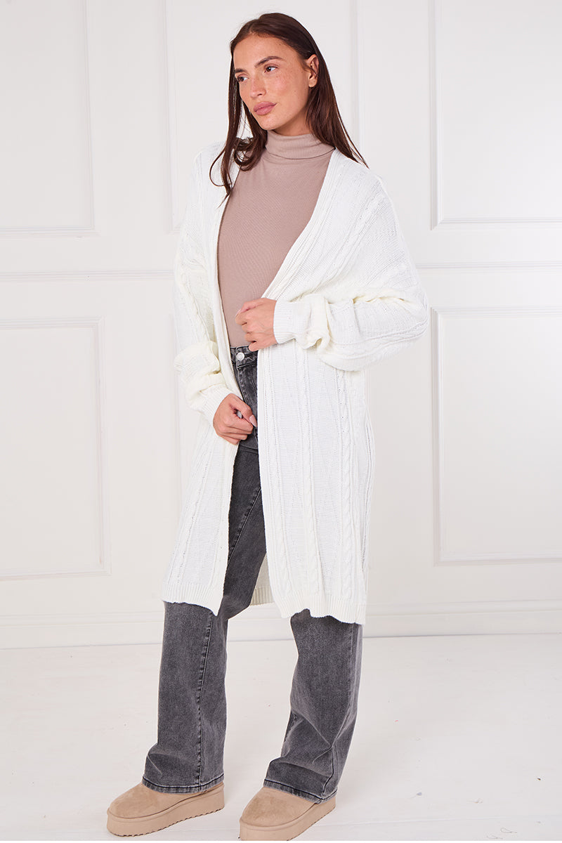 Woman wearing a white cardigan, beige turtleneck, and dark jeans against a white background