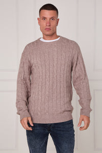 Man wearing a beige cable knit sweater and dark jeans against a white background