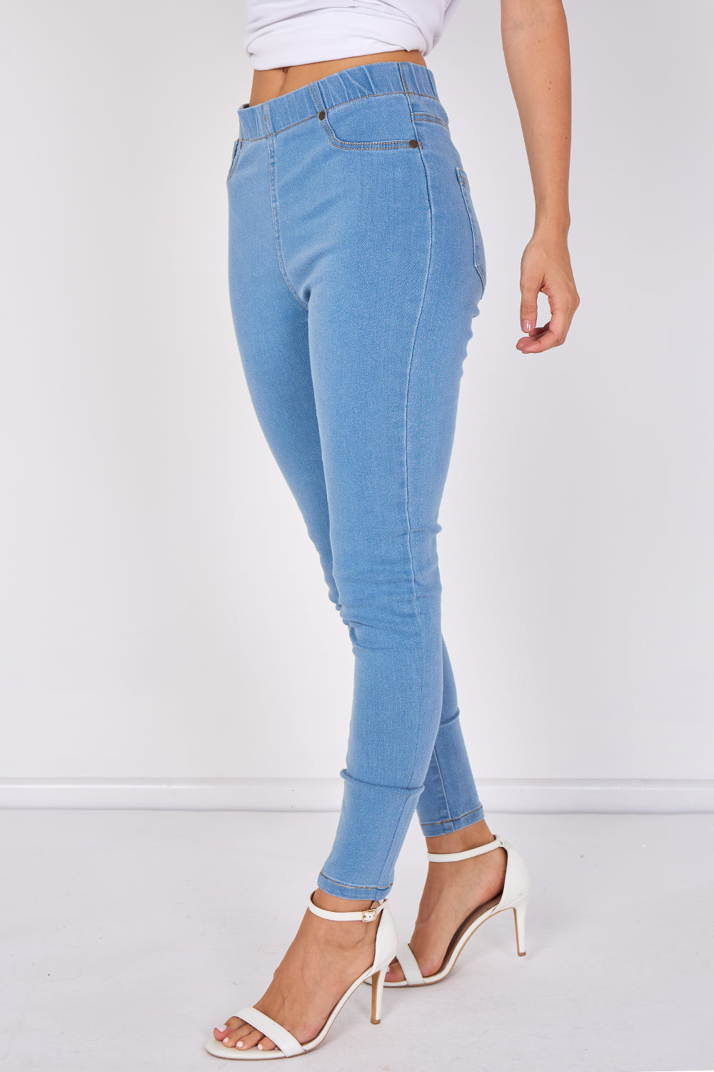 A model wearing light blue skinny jeans with a high-waisted, elastic waistband, paired with white strappy high-heeled sandals against a white studio background.