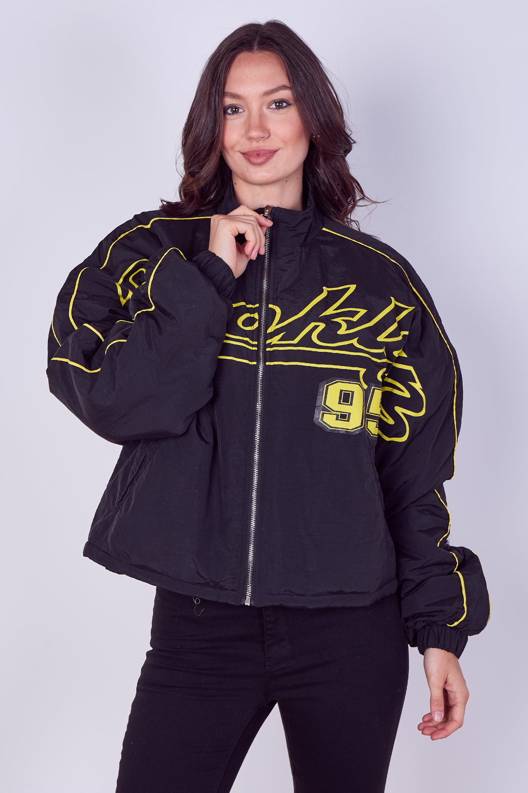 A woman modeling a black zip-up jacket with yellow graphic lettering and the number 95 on the front, paired with black pants, against a light gray studio background.