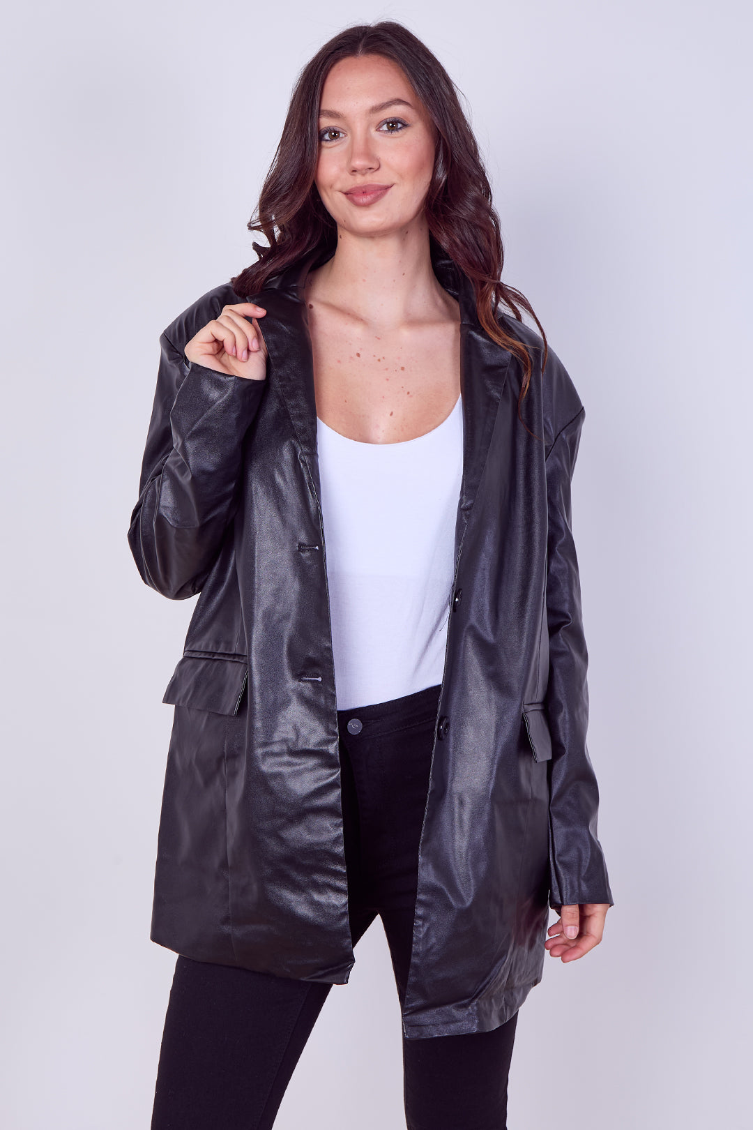 A woman modeling a black faux-leather jacket over a white tank top and black pants, standing against a light gray studio background.