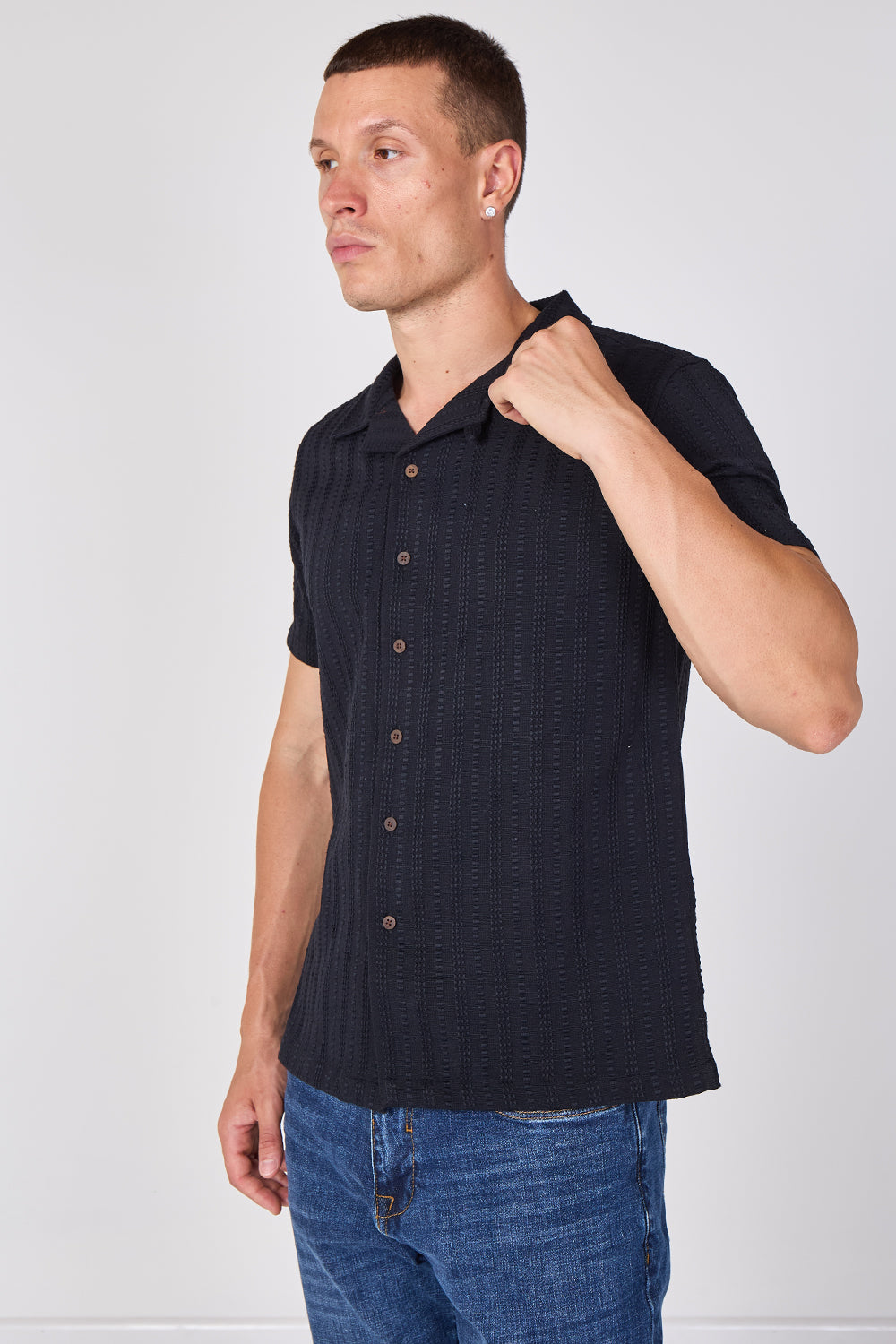 A man wearing a dark navy blue short-sleeve button-up shirt with a subtle textured pattern, paired with blue jeans, photographed against a light gray studio background.