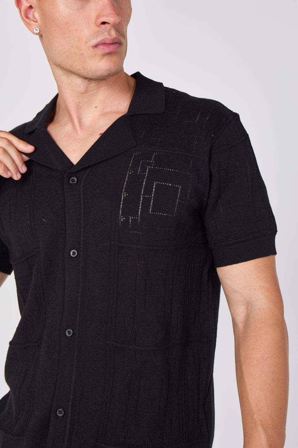 A close-up of a man wearing a black short-sleeve button-up shirt with a textured, vertically ribbed fabric and geometric stitched details on the chest pocket.