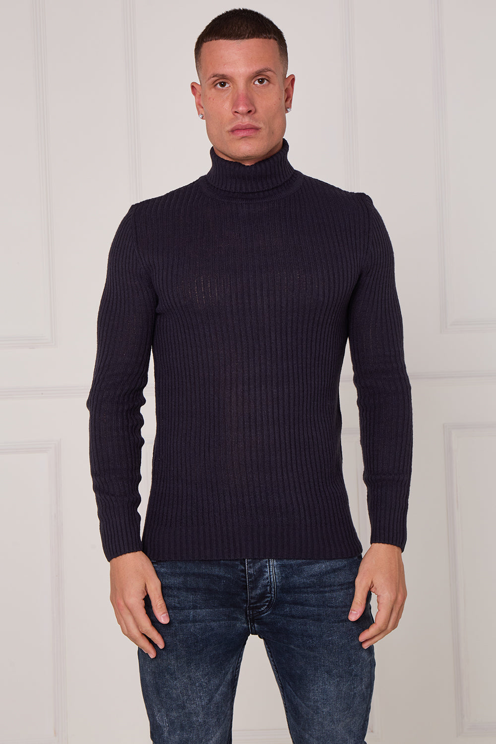 Man wearing a dark ribbed turtleneck sweater and jeans against a white wall.