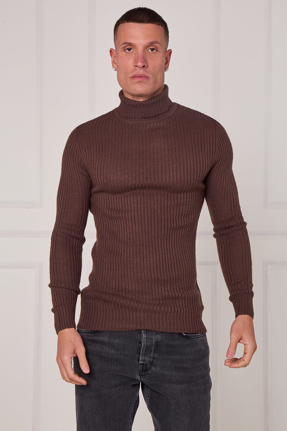 Man wearing a brown ribbed turtleneck sweater and dark jeans against a white background