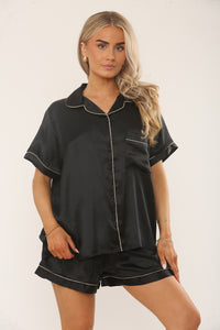 Woman wearing a black satin pajama set with white piping on a plain background