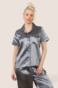 Woman wearing a grey satin pyjama set with short sleeves and long trousers, featuring dark-coloured piping details, standing against a neutral background