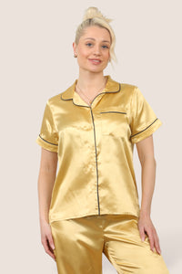Women’s gold satin short sleeve pajama set with button-up top and full-length pants, worn by a model standing barefoot. Lightweight 100% polyester sleepwear with black piping details, ideal for loungewear and bedtime comfort.