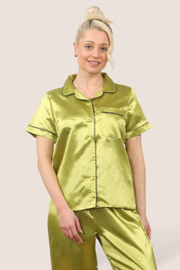 Woman wearing a shiny green satin pajama set with short sleeves and long pants, standing barefoot against a neutral background
