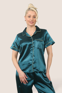 Woman wearing a teal satin pyjama set with short sleeves and long trousers, featuring light-coloured piping details, standing against a neutral background