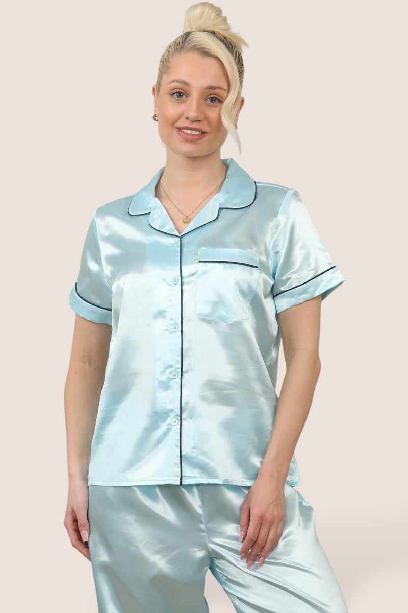 Woman wearing a light blue satin pyjama set with short sleeves and long trousers, featuring dark-coloured piping details, standing against a neutral background