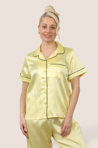 Woman wearing a light yellow satin pyjama set with short sleeves and long trousers, featuring dark-coloured piping details, standing against a neutral background