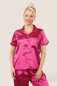 Woman wearing a hot pink satin pyjama set with short sleeves and long trousers, featuring dark-coloured piping details, standing against a neutral background