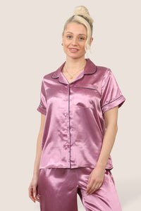 Woman wearing a dusty pink satin pyjama set with short sleeves and long trousers, featuring dark-coloured piping details, standing against a neutral background