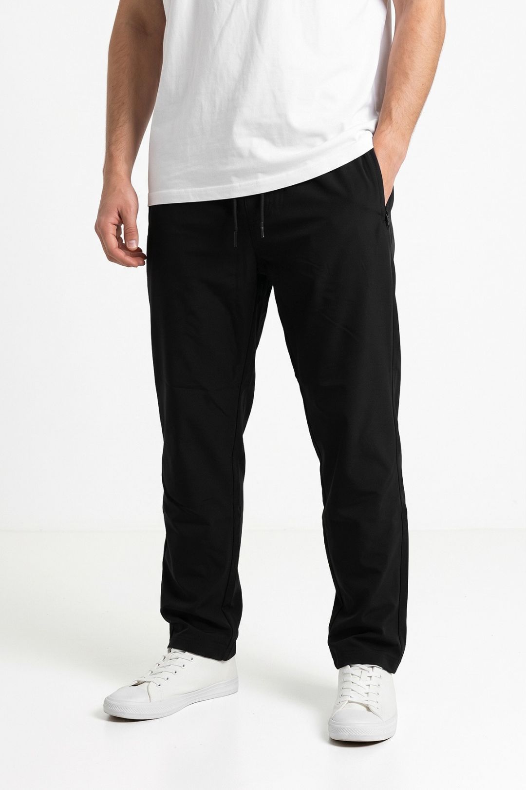 A person wearing black drawstring trousers with a white t-shirt and white sneakers, shown from the chest down against a clean white studio background.