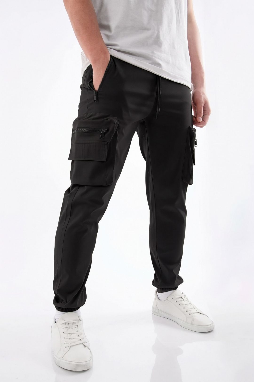 A person wearing black cargo jogger pants with side flap pockets, paired with a white t-shirt and white sneakers, shown from the waist down on a white studio background.